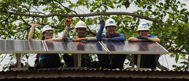4 people sit on behind solar panels on tower roof smiling with thumbs up