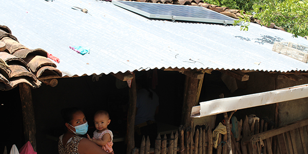 Masked woman holding a baby on a rural home with a solar panel on the roof
