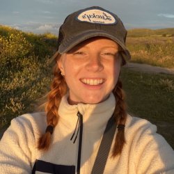 Smiling selfie of Emma at dusk in a field. They wear a baseball cap with a Lucky's logo, their auburn shoulder-length hair is tied into 2 braids.