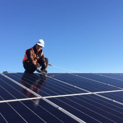 A trainee wearing an orange vest smiles while securing a solar panel on top of a roof. The sky is blue behind him.
