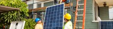 group of people in hard hats on the ground and on the roof of a house raising a solar panel from the ground up onto the roof of a house