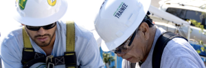 close up of two men installing solar on a roof wear white hard hats and long sleeved white GRID shirts