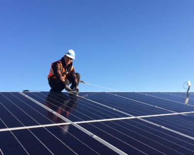 A trainee wearing an orange vest smiles while securing a solar panel on top of a roof. The sky is blue behind him.