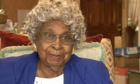 Elderly woman with gray curly hair and glasses wearing blue, seated in home setting