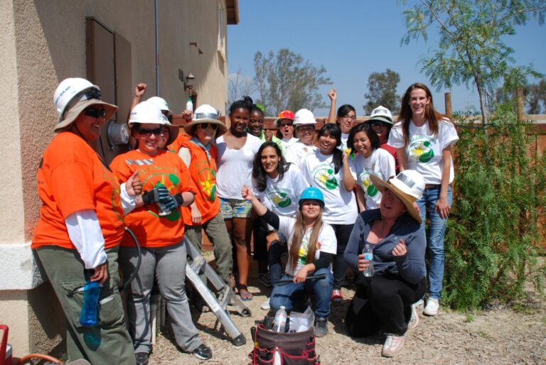 Group of volunteers in hard hats and orange/white shirts celebrating Women's History Month at a construction site