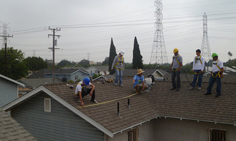 Construction workers installing roofing materials on a residential house with power lines and transmission towers visible in the background