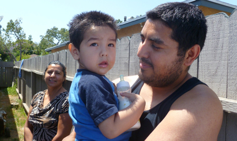 Man holding toddler in blue shirt outdoors near house, woman standing in background