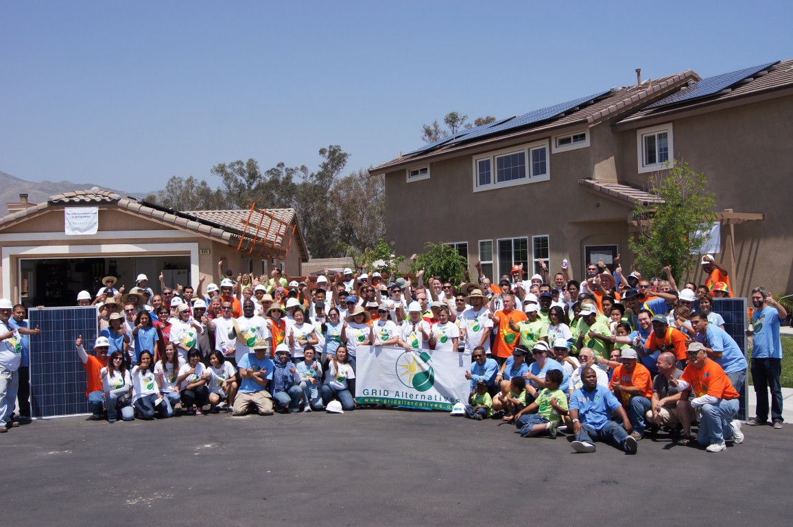 Large group of volunteers in orange and blue shirts holding GRID Alternatives banner at solar installation event
