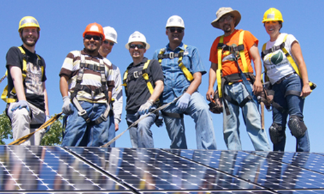 Seven construction workers in hard hats and safety gear stand on solar panels under blue sky