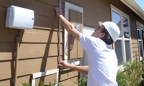 Worker in white hard hat and shirt installing electrical panel on house exterior