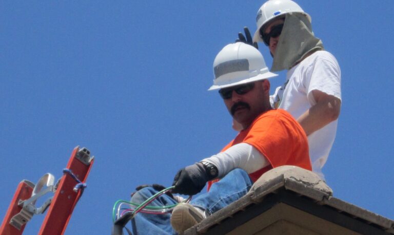 Two construction workers on a roof against a clear blue sky, one wearing an orange shirt and black gloves working with tools