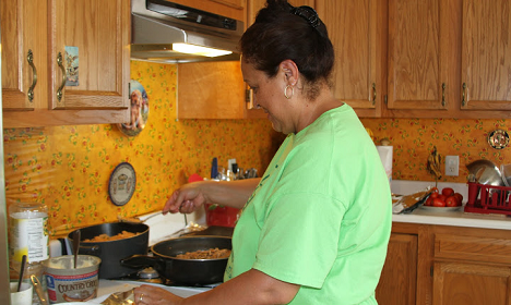 Woman in green shirt cooking at stove in kitchen with yellow tile backsplash