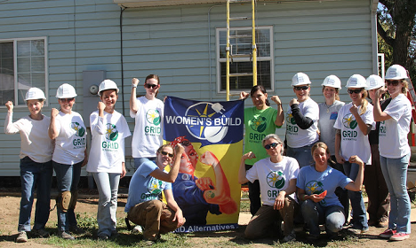 Group of volunteers in hard hats and matching shirts holding a "Women Build" banner in front of a house