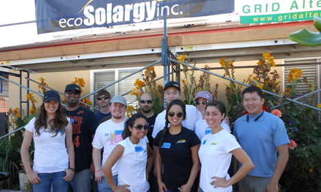Group of people in white shirts posing at EcoSolargy Inc. facility with dried plants and "GRID Alte" signage visible