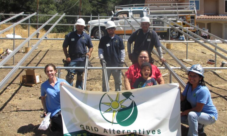 GRID Alternatives team and community members holding banner at solar installation site with workers and equipment in background
