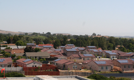 Suburban neighborhood with numerous homes featuring solar panels on red-tiled roofs, green trees, and mountains in background