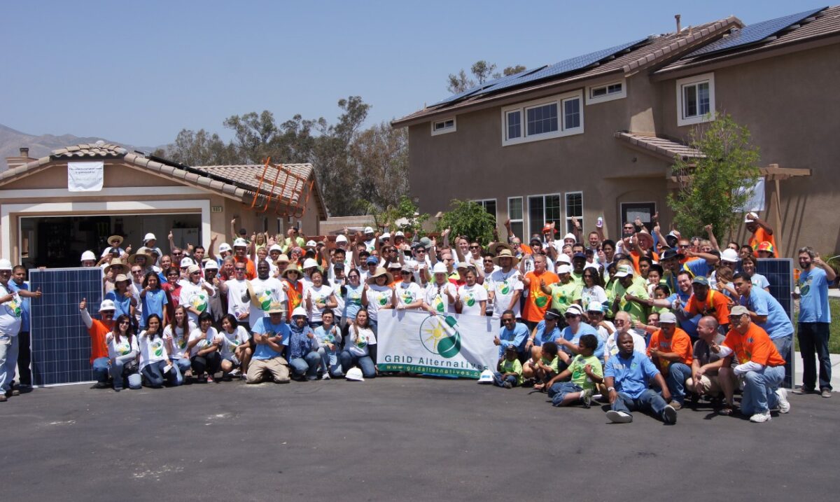 Large group of volunteers in safety vests and hard hats gathered in driveway holding GRID Alternatives banner in front of solar-installed house