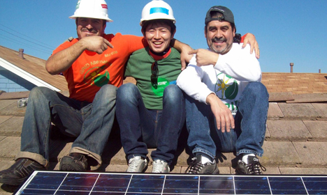 Three men in hard hats and safety gear smile while sitting on a solar panel installation