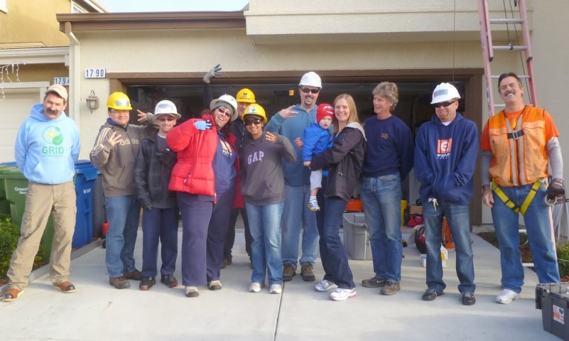 Group of volunteers wearing hard hats and safety gear posing together at a garage during a community work project