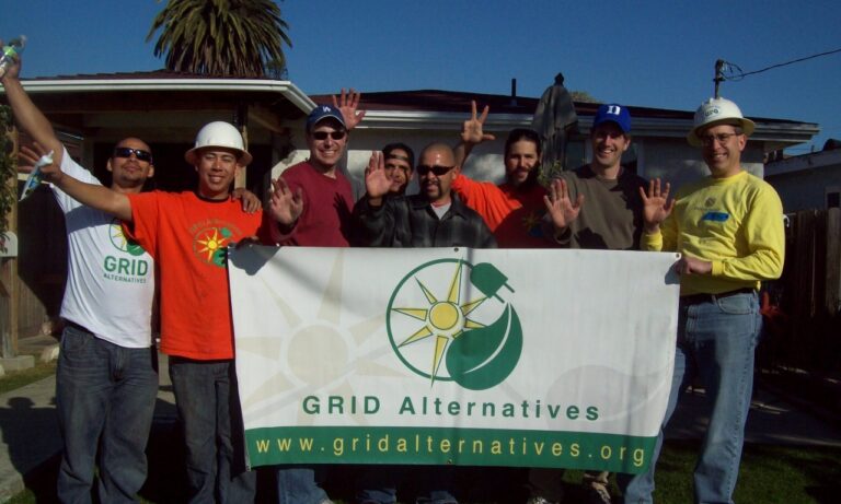 Nine people holding GRID Alternatives banner in front of house with solar panels