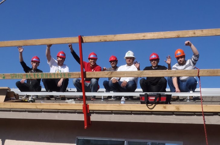 Eight construction workers wearing hard hats celebrate on a wooden beam during a solar installation project