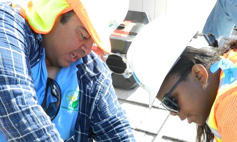 Two young interns in safety gear and hard hats examine solar panel equipment during a work training session