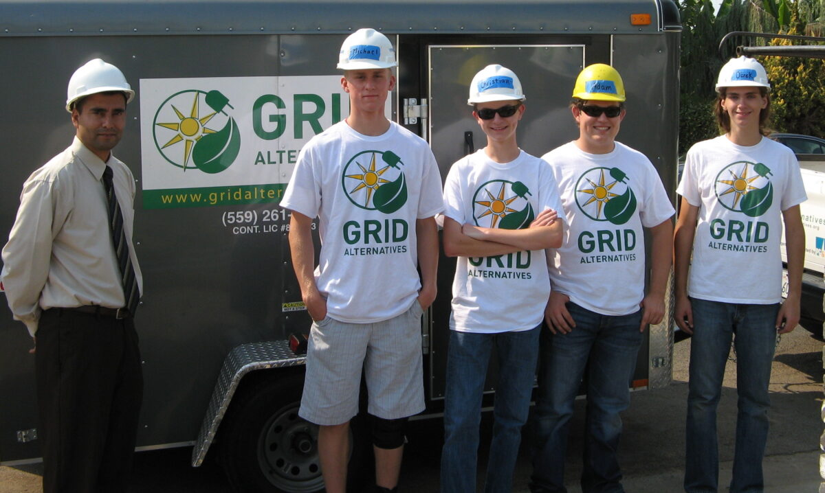 Six people in hard hats stand in front of a GRID Alternatives trailer, wearing matching white GRID Alternatives t-shirts with a sun and leaf logo