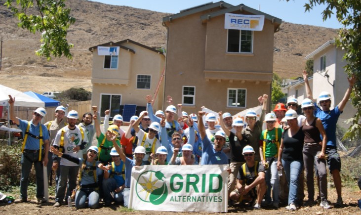 Large group of people wearing hard hats and safety vests holding a GRID Alternatives banner in front of a building under construction