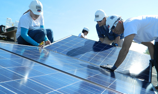 Workers in white hard hats installing solar panels on a rooftop under blue sky