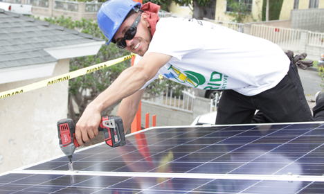 Worker installing solar panels on rooftop with power drill and caution tape visible