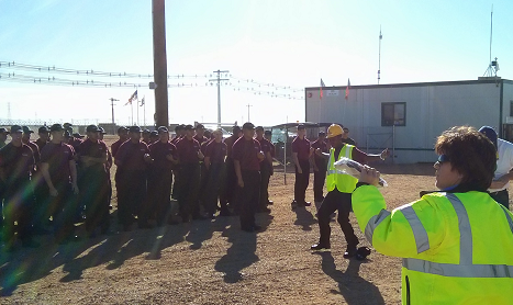 Large group gathered outdoors near industrial buildings with safety personnel in high-visibility vests directing the crowd