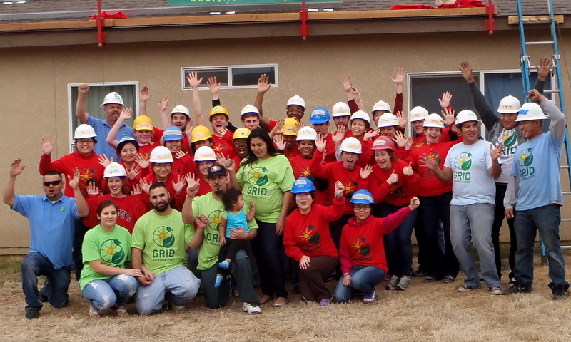 Large group of volunteers in hard hats and red or green GRID shirts raising hands in front of building under construction