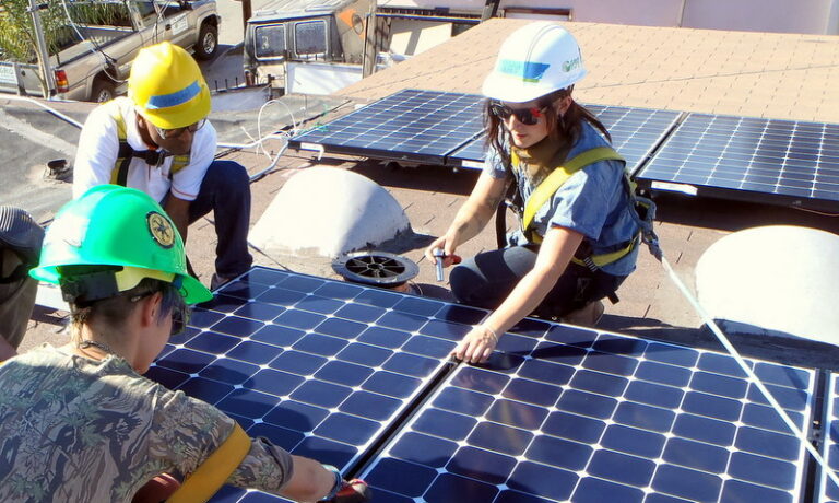 Three workers installing solar panels on a roof with safety equipment