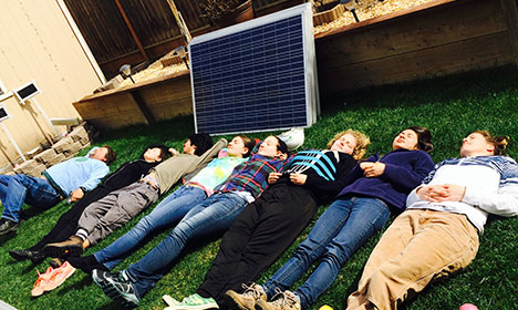 Group of people lying on grass beneath a solar panel during an educational solar energy workshop