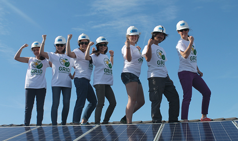 Seven people in white GRi shirts and hard hats stand on solar panels, smiling and celebrating with fists raised