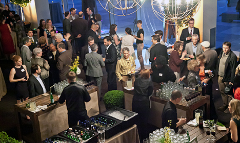 Formal event with guests in business attire mingling around cocktail tables and floral arrangements indoors