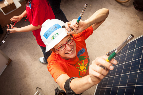 Person in orange shirt and white hard hat holding solar panel and tools, smiling at camera