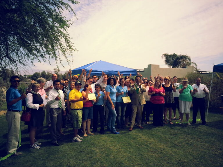 Large group celebrating outdoors under blue tent with thumbs up gestures, green lawn and buildings in background
