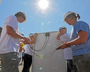 Multiple volunteers in hard hats installing solar panel equipment outdoors under bright sunlight