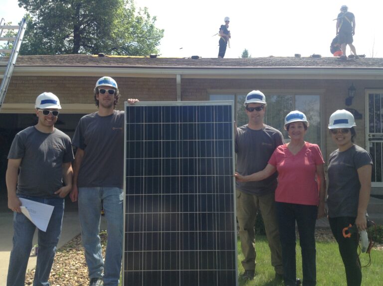 Six workers in hard hats and safety gear display a solar panel in front of a house during installation