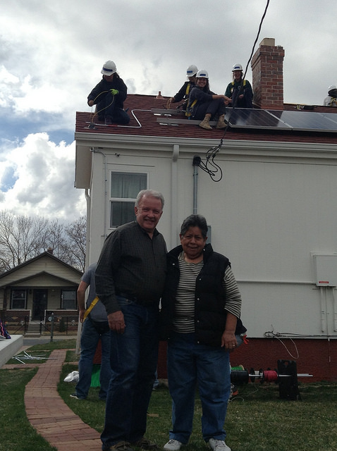 Two homeowners smile in front of their house while workers install a new roof above
