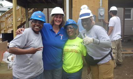 Four volunteers wearing hard hats pose together at a construction site during a community rebuild project