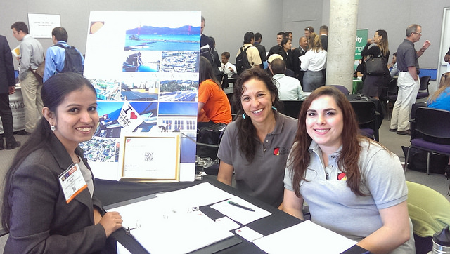 Three women smile at a table displaying solar energy project photos and materials at a jobs fair event