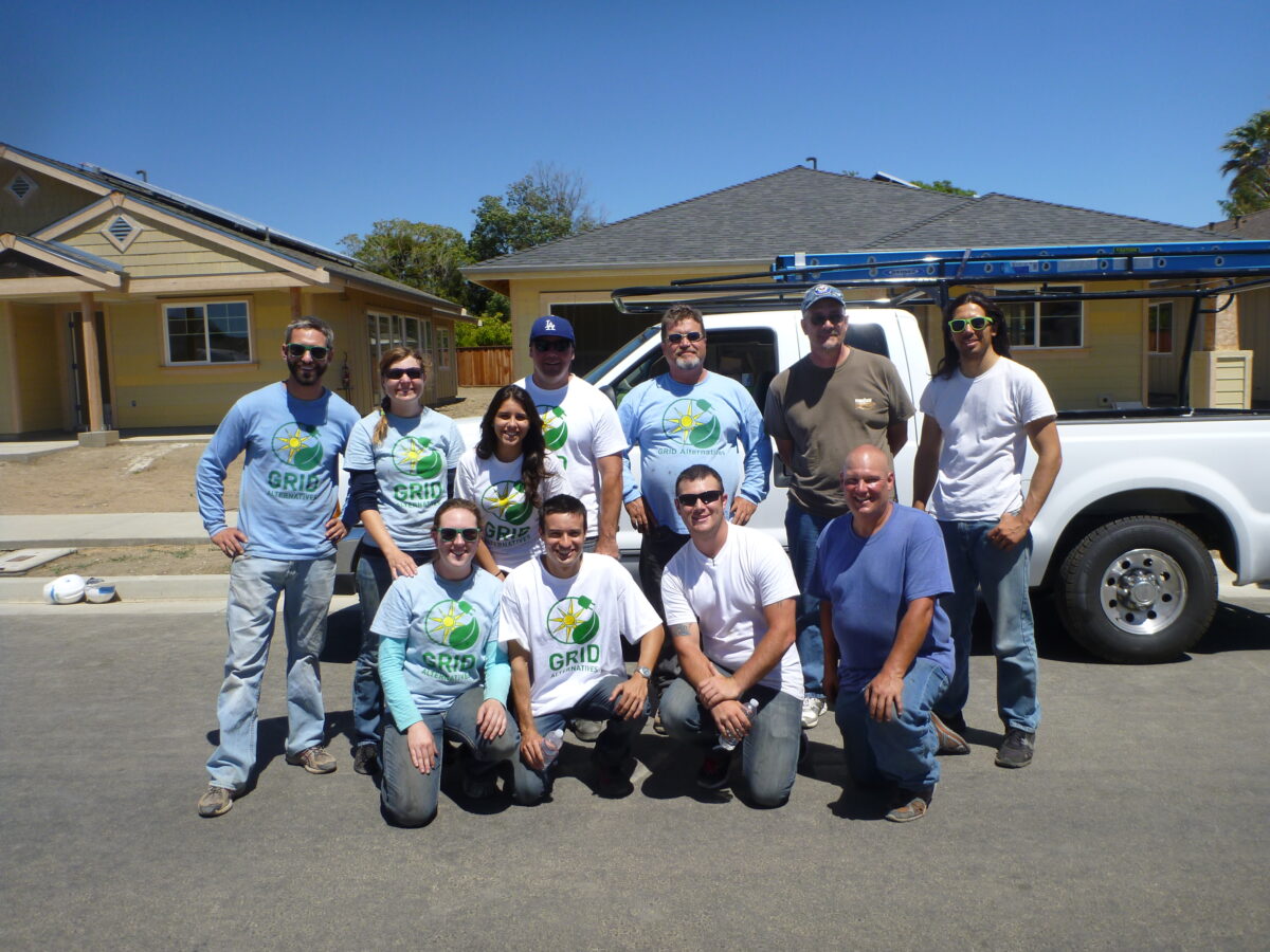 Volunteer team in GRID t-shirts poses in front of yellow house with white pickup truck on sunny day