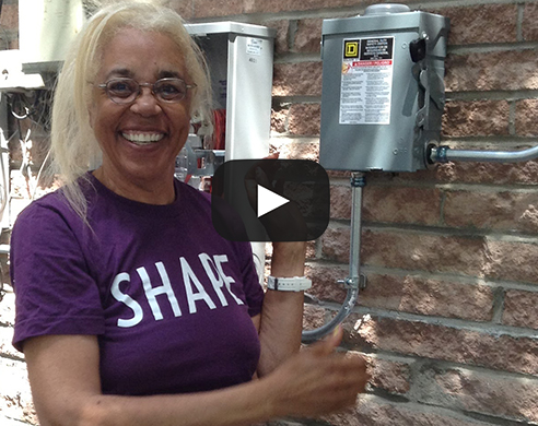 Woman in purple "Share" shirt smiling next to electrical panel on brick wall