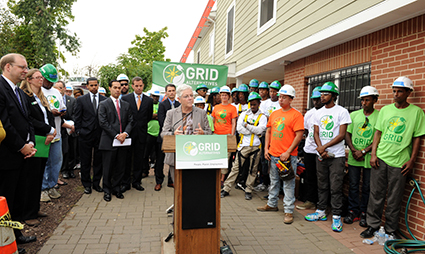 Officials and youth in green shirts at GRID podium during solar policy event for low-income families
