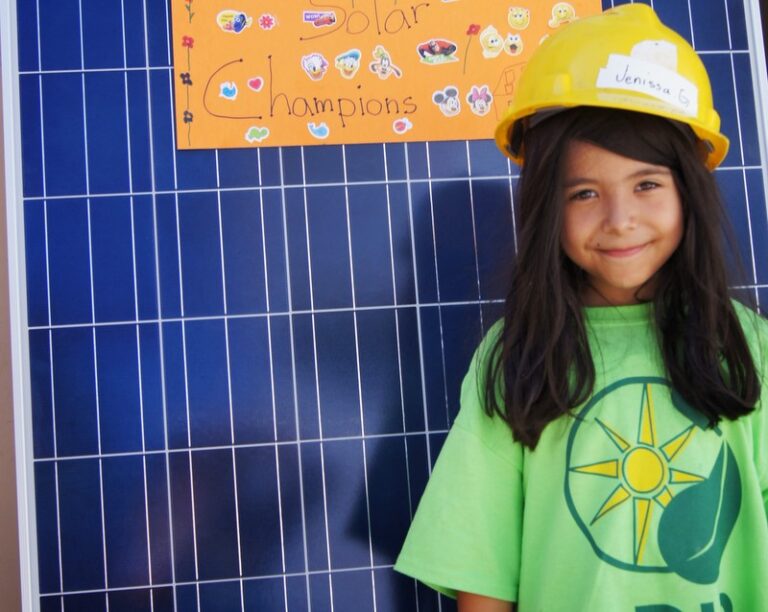 Child in green solar shirt and yellow hard hat standing before solar panel with "Solar Champions" poster