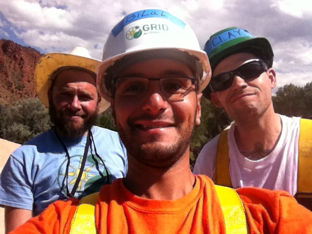 Three construction workers wearing hard hats and safety vests smile at the camera outdoors