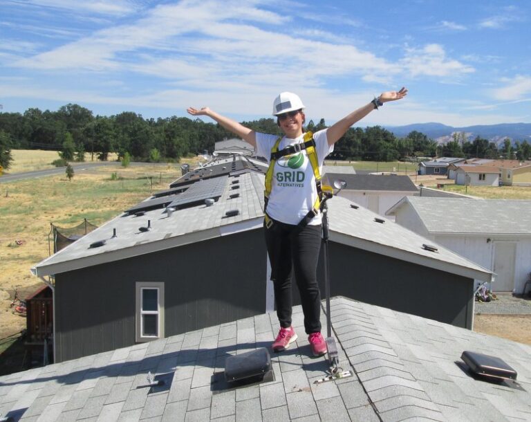 Worker celebrating on residential rooftop with solar panels installed, wearing GRID Alternatives safety gear