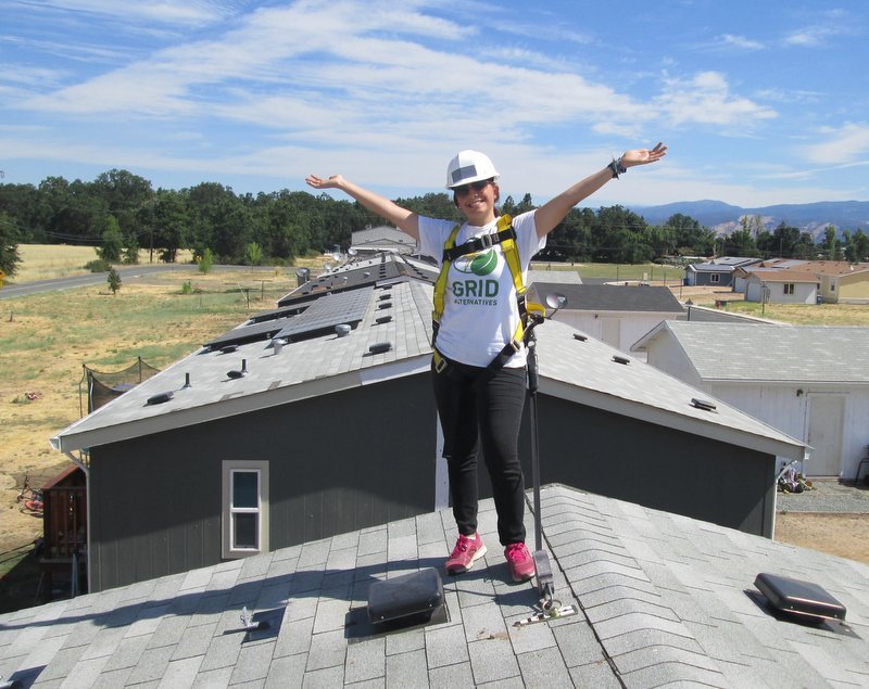 Worker celebrating on residential rooftop with solar panels installed, wearing GRID Alternatives safety gear